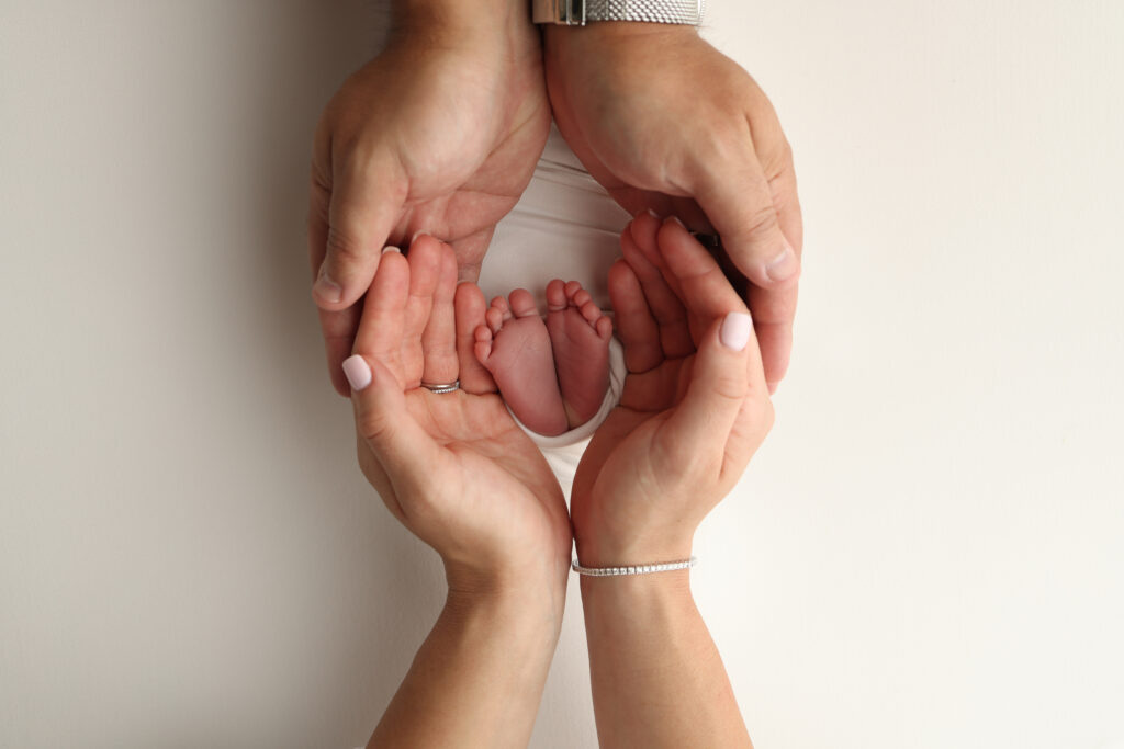 The palms of the parents. A father and mother hold the feet of a newborn child in a white blanket on a white background.. The feet of a newborn in the hands of parents. Photo of foot, heels and toes.